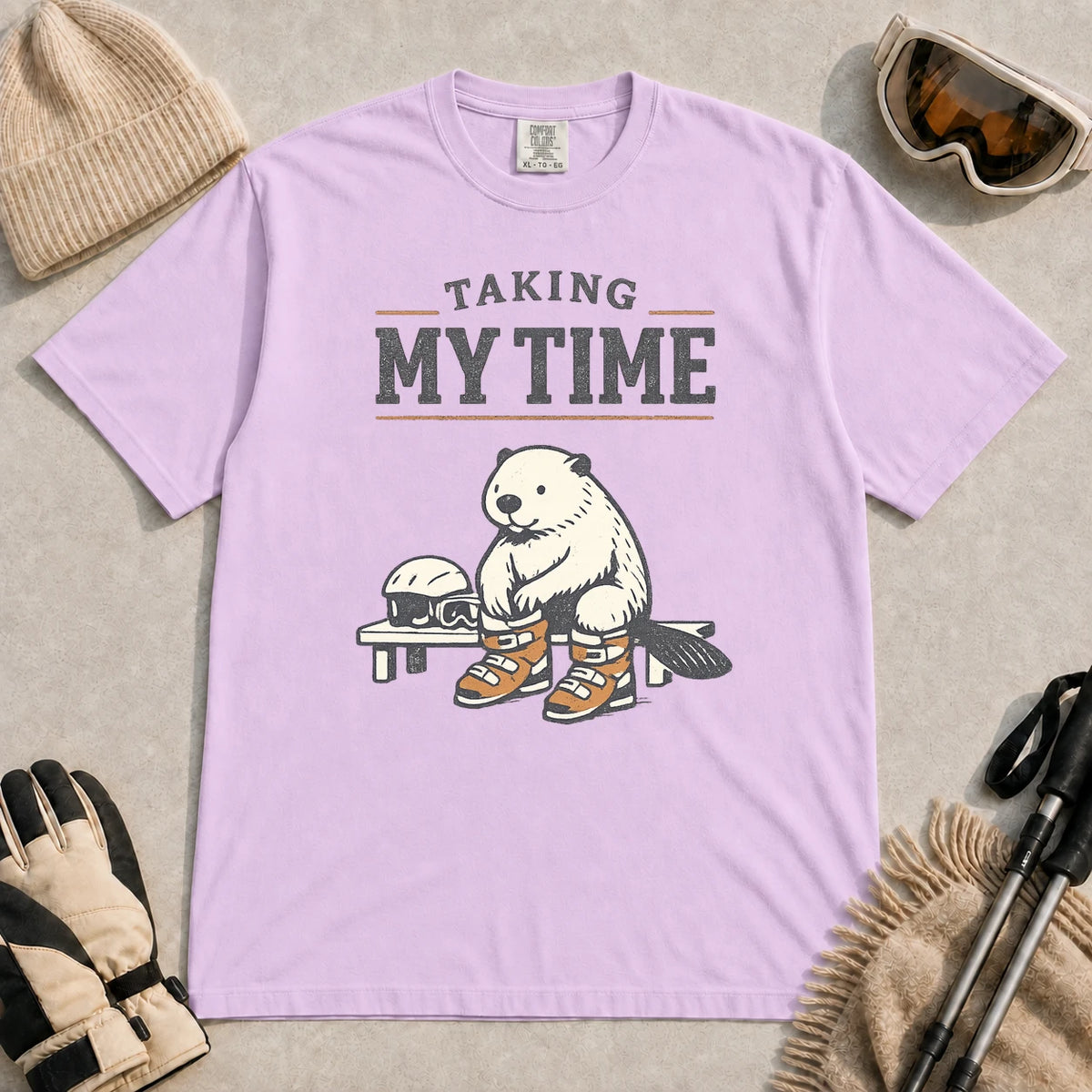 Orchid tee featuring a relaxed beaver sitting on a bench in ski boots beside a helmet and goggles, with the phrase “Taking My Time.”