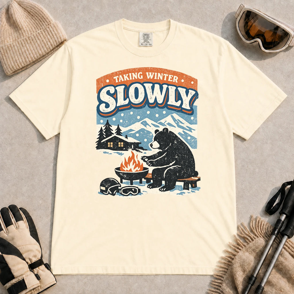 Ivory tee featuring a relaxed bear warming up beside a fire in a snowy mountain setting, with the phrase “Taking Winter Slowly.”