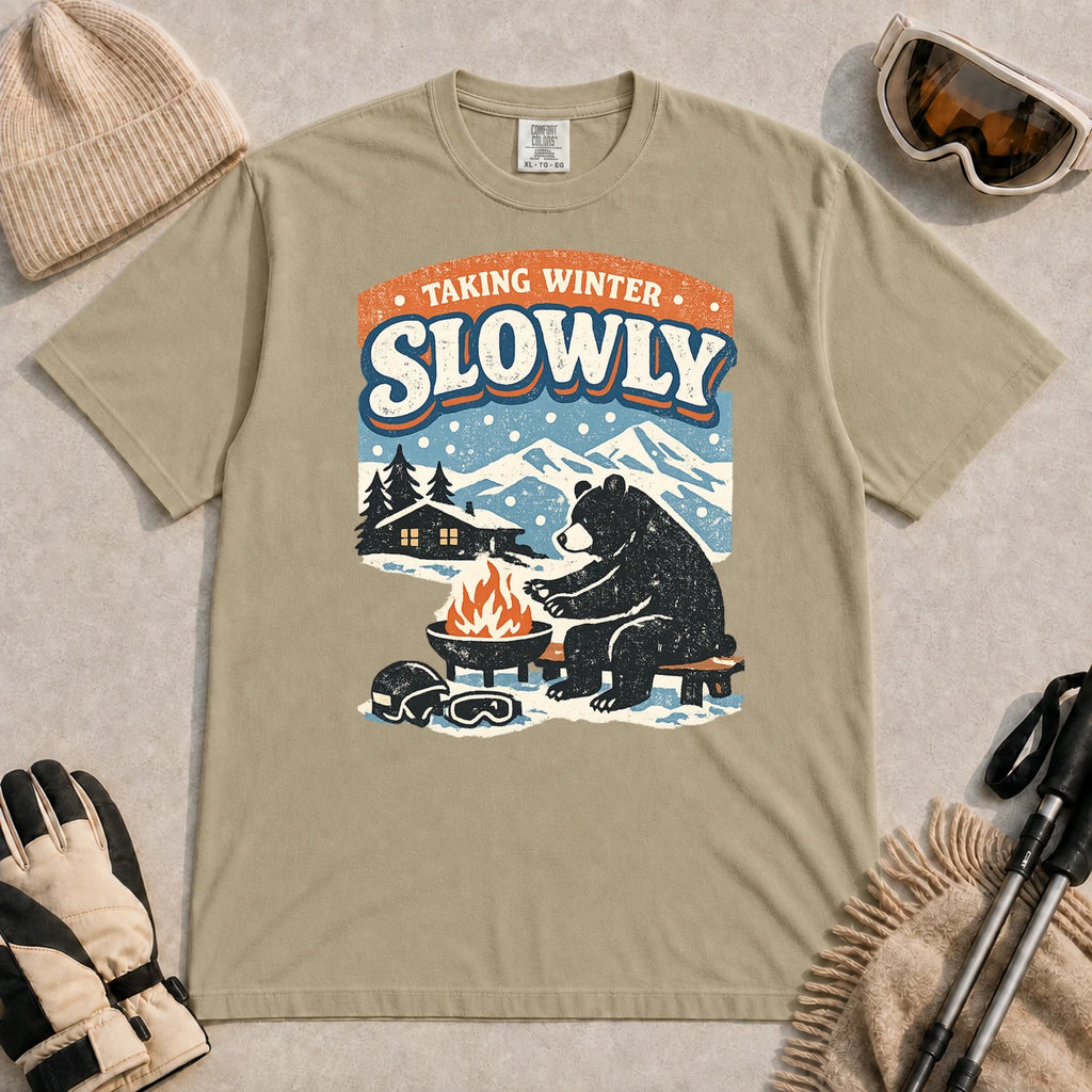 Khaki tee featuring a relaxed bear warming up beside a fire in a snowy mountain setting, with the phrase “Taking Winter Slowly.”