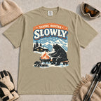Khaki tee featuring a relaxed bear warming up beside a fire in a snowy mountain setting, with the phrase “Taking Winter Slowly.”