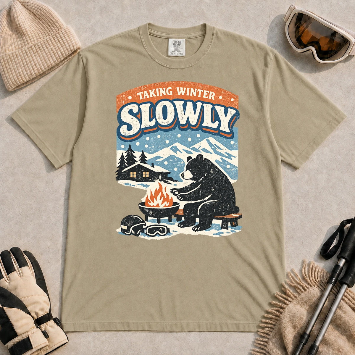 Khaki tee featuring a relaxed bear warming up beside a fire in a snowy mountain setting, with the phrase “Taking Winter Slowly.”