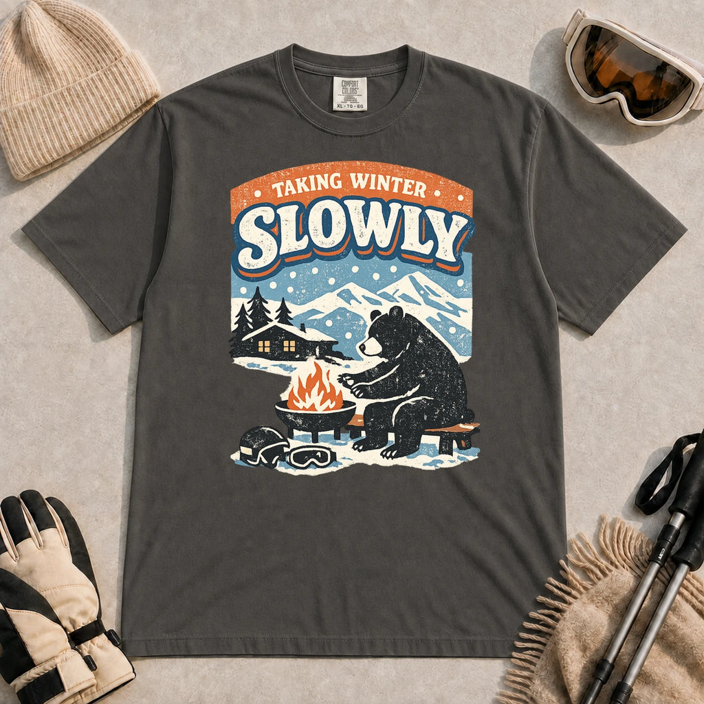 Pepper tee featuring a relaxed bear warming up beside a fire in a snowy mountain setting, with the phrase “Taking Winter Slowly.”