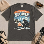 Pepper tee featuring a relaxed bear warming up beside a fire in a snowy mountain setting, with the phrase “Taking Winter Slowly.”