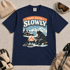 True Navy tee featuring a relaxed bear warming up beside a fire in a snowy mountain setting, with the phrase “Taking Winter Slowly.”