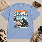 Washed Denim tee featuring a relaxed bear warming up beside a fire in a snowy mountain setting, with the phrase “Taking Winter Slowly.”