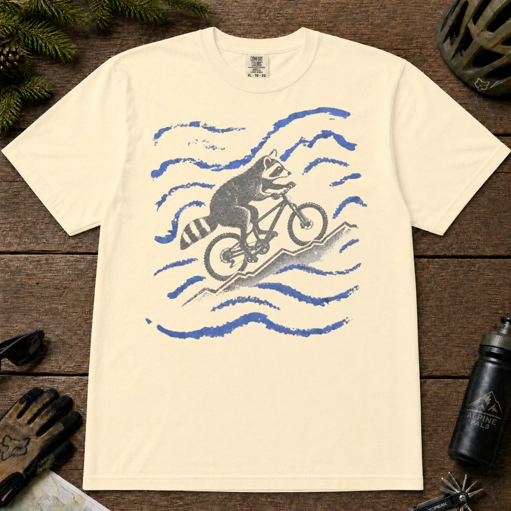 Ivory tee featuring a raccoon mountain biking uphill through a playful blue wavy mountain scene.