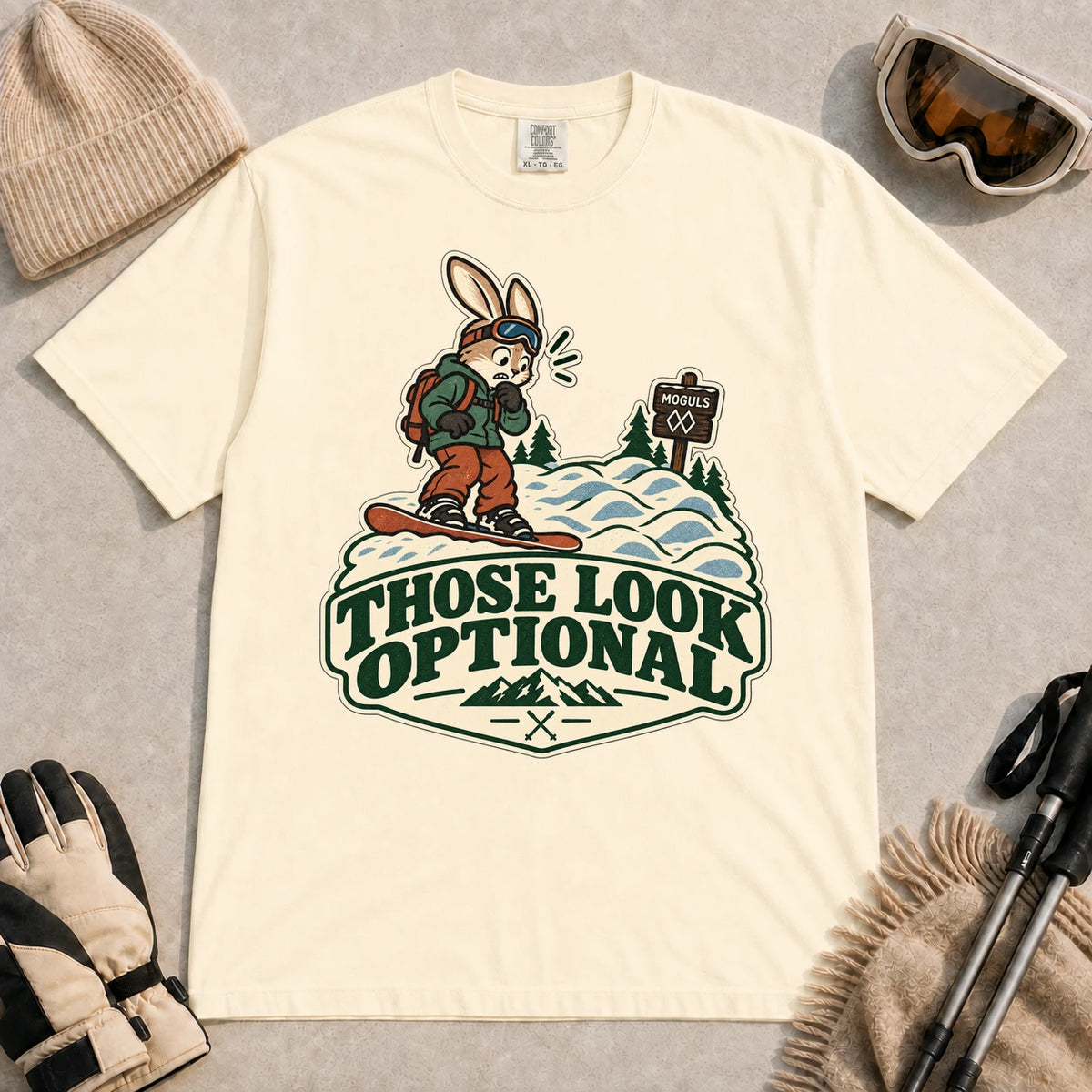 Ivory tee featuring a cautious bunny snowboarder facing a mogul run beside a “Moguls” sign above the phrase “Those Look Optional.”