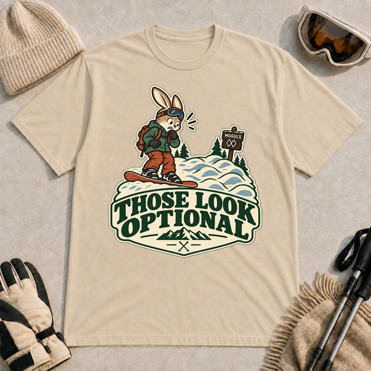 Sand tee featuring a cautious bunny snowboarder facing a mogul run beside a “Moguls” sign above the phrase “Those Look Optional.”