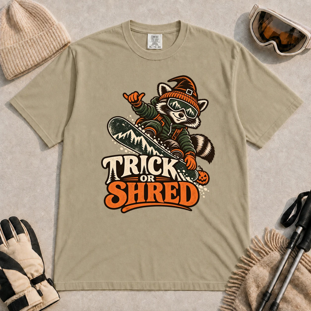 Khaki tee featuring a raccoon snowboarding in Halloween gear and a witch hat above the phrase “Trick or Shred.”