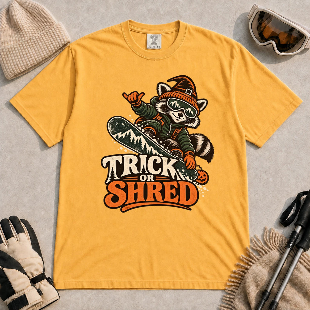 Mustard tee featuring a raccoon snowboarding in Halloween gear and a witch hat above the phrase “Trick or Shred.”