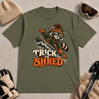 Military Green tee featuring a raccoon snowboarding in Halloween gear and a witch hat above the phrase “Trick or Shred.”