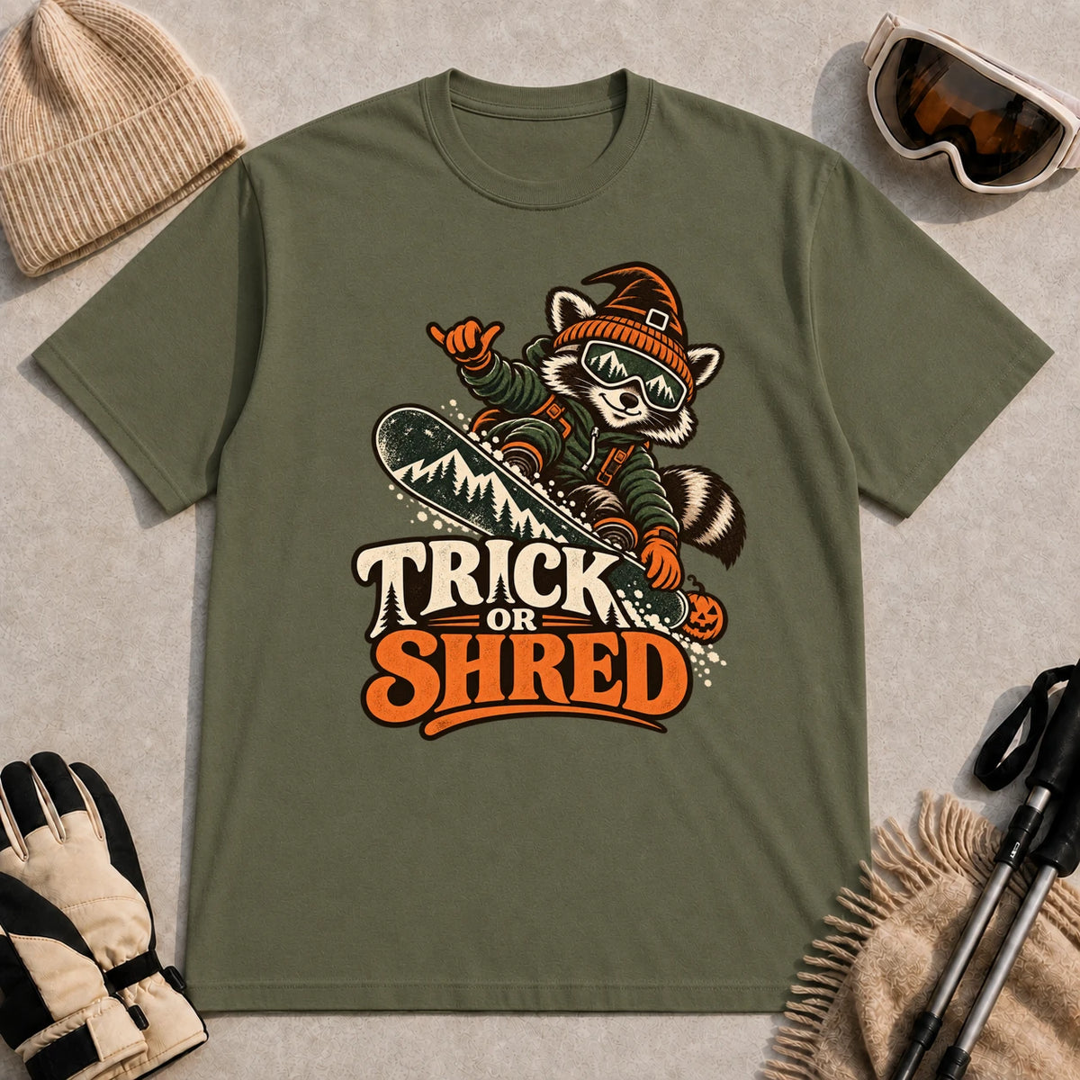 Military Green tee featuring a raccoon snowboarding in Halloween gear and a witch hat above the phrase “Trick or Shred.”