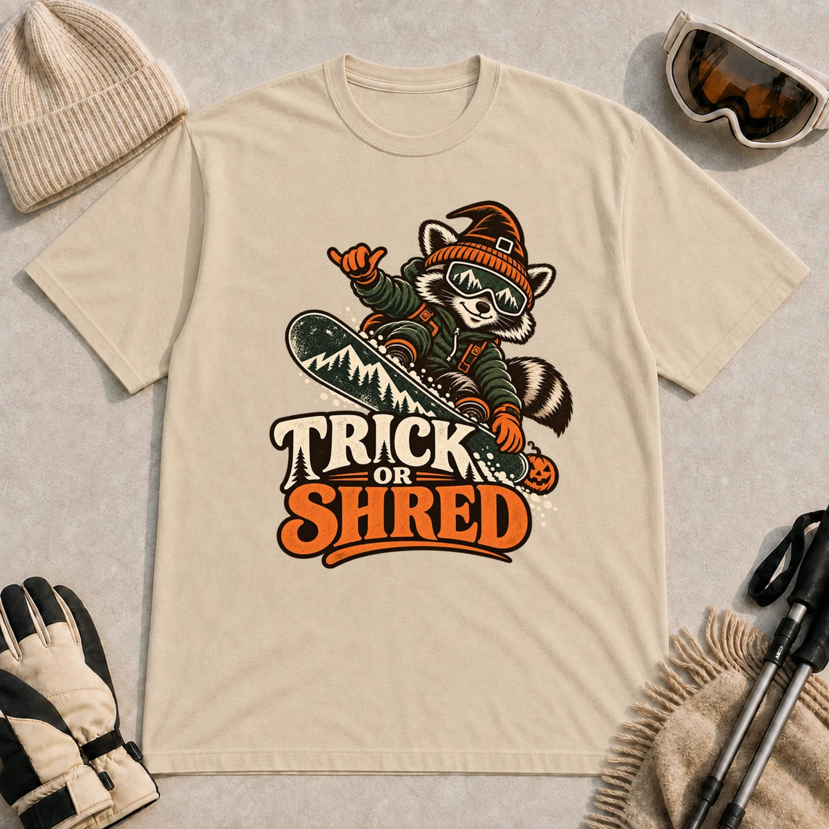 Sand tee featuring a raccoon snowboarding in Halloween gear and a witch hat above the phrase “Trick or Shred.”