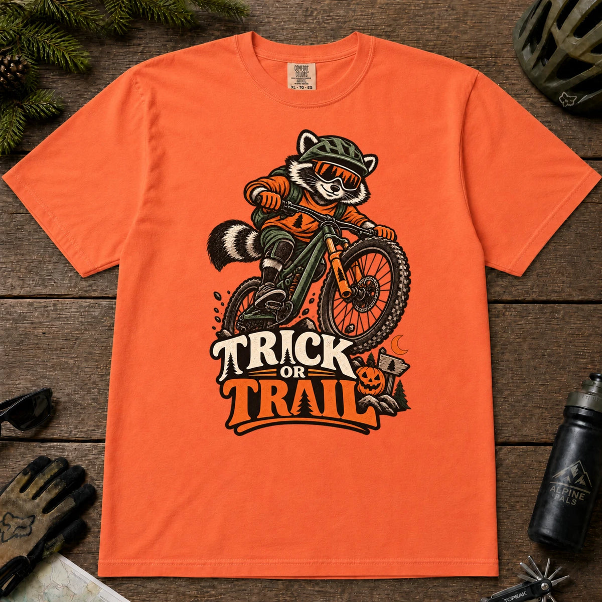Burnt Orange tee featuring a raccoon mountain biking in Halloween gear above a pumpkin and trail sign, with the phrase “Trick or Trail.”