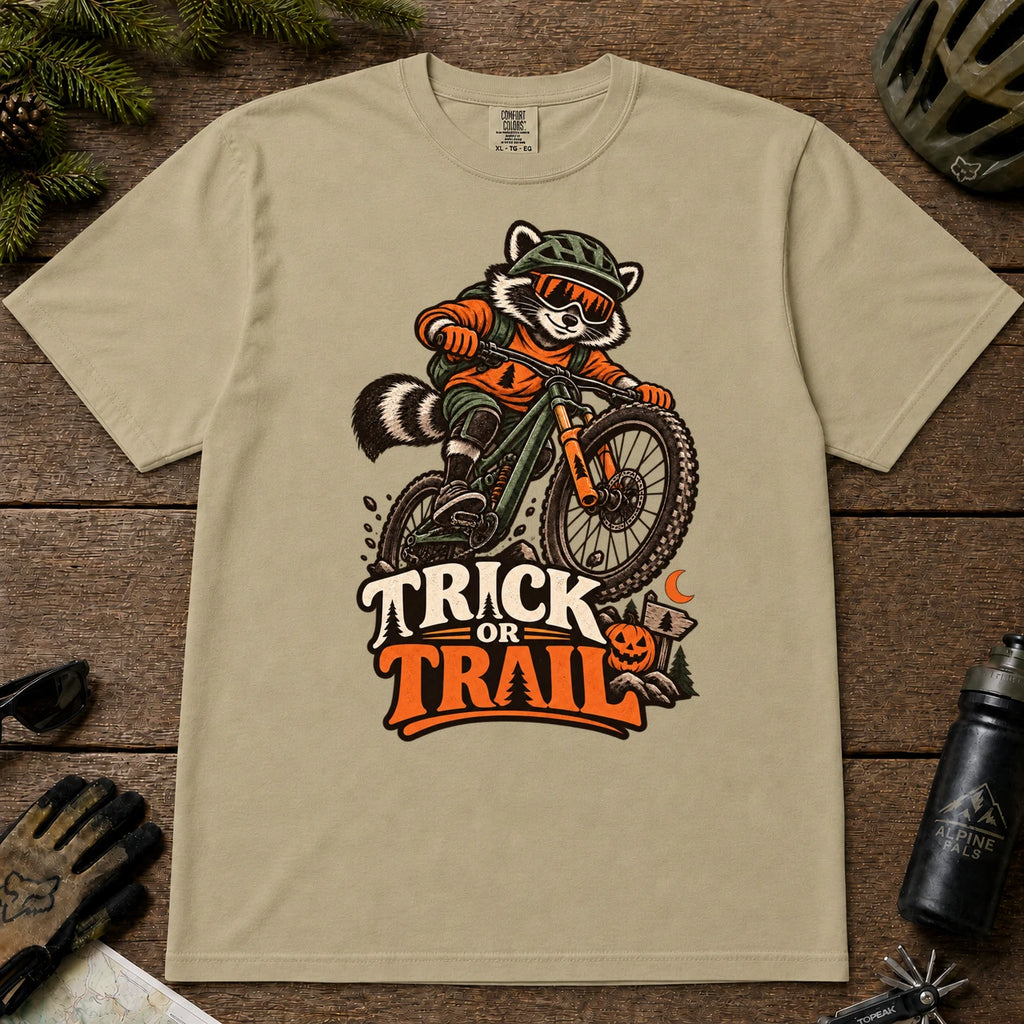 Khaki tee featuring a raccoon mountain biking in Halloween gear above a pumpkin and trail sign, with the phrase “Trick or Trail.”
