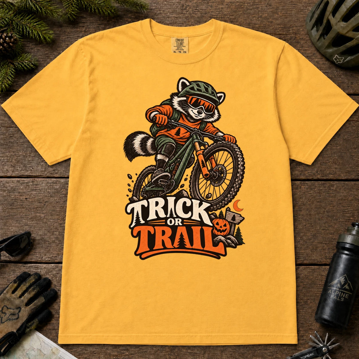 Mustard tee featuring a raccoon mountain biking in Halloween gear above a pumpkin and trail sign, with the phrase “Trick or Trail.”