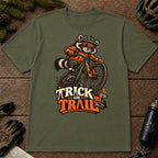 Military Green tee featuring a raccoon mountain biking in Halloween gear above a pumpkin and trail sign, with the phrase “Trick or Trail.”