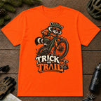 Orange tee featuring a raccoon mountain biking in Halloween gear above a pumpkin and trail sign, with the phrase “Trick or Trail.”