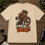 Sand tee featuring a raccoon mountain biking in Halloween gear above a pumpkin and trail sign, with the phrase “Trick or Trail.”