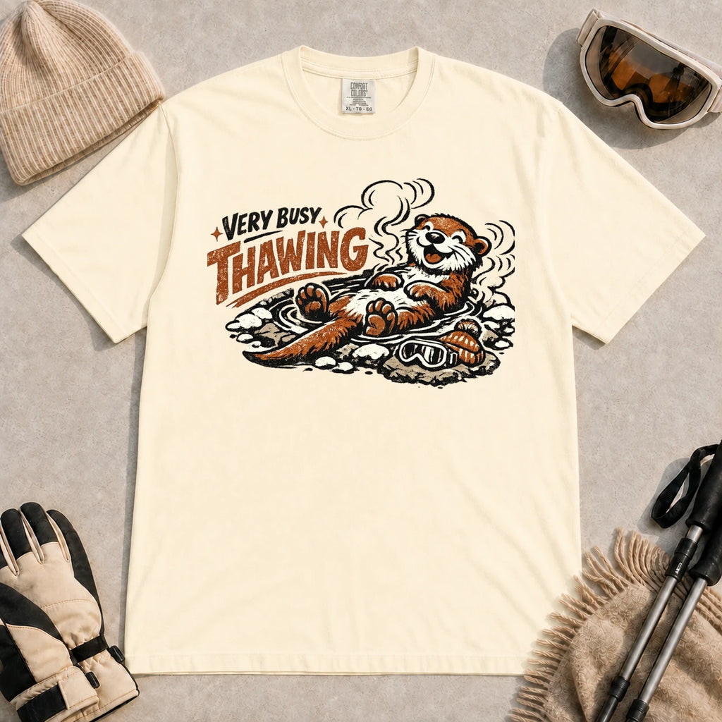 Ivory tee featuring a relaxed otter thawing in a warm mountain hot spring beside ski goggles and a beanie, with the phrase “Very Busy Thawing.”