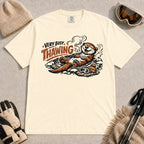 Ivory tee featuring a relaxed otter thawing in a warm mountain hot spring beside ski goggles and a beanie, with the phrase “Very Busy Thawing.”