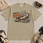 Khaki tee featuring a relaxed otter thawing in a warm mountain hot spring beside ski goggles and a beanie, with the phrase “Very Busy Thawing.”