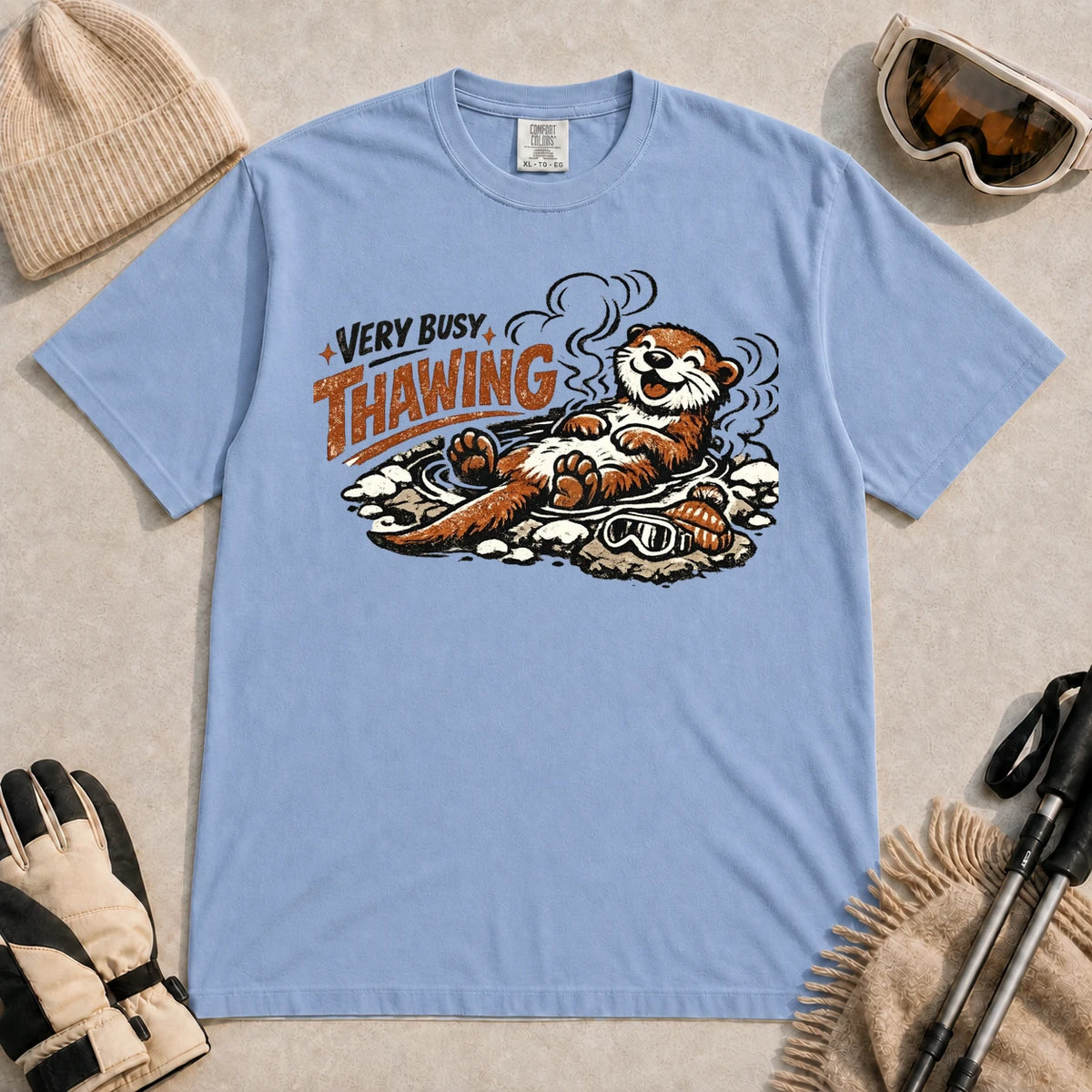 Washed Denim tee featuring a relaxed otter thawing in a warm mountain hot spring beside ski goggles and a beanie, with the phrase “Very Busy Thawing.”