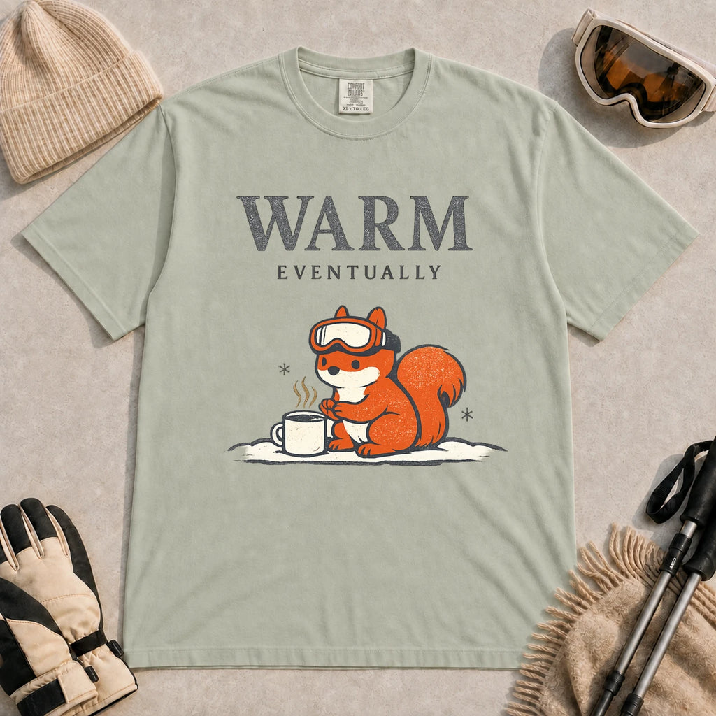 Bay tee featuring a chilly squirrel in ski goggles warming its paws beside a steaming mug in the snow, with the phrase “Warm Eventually.”