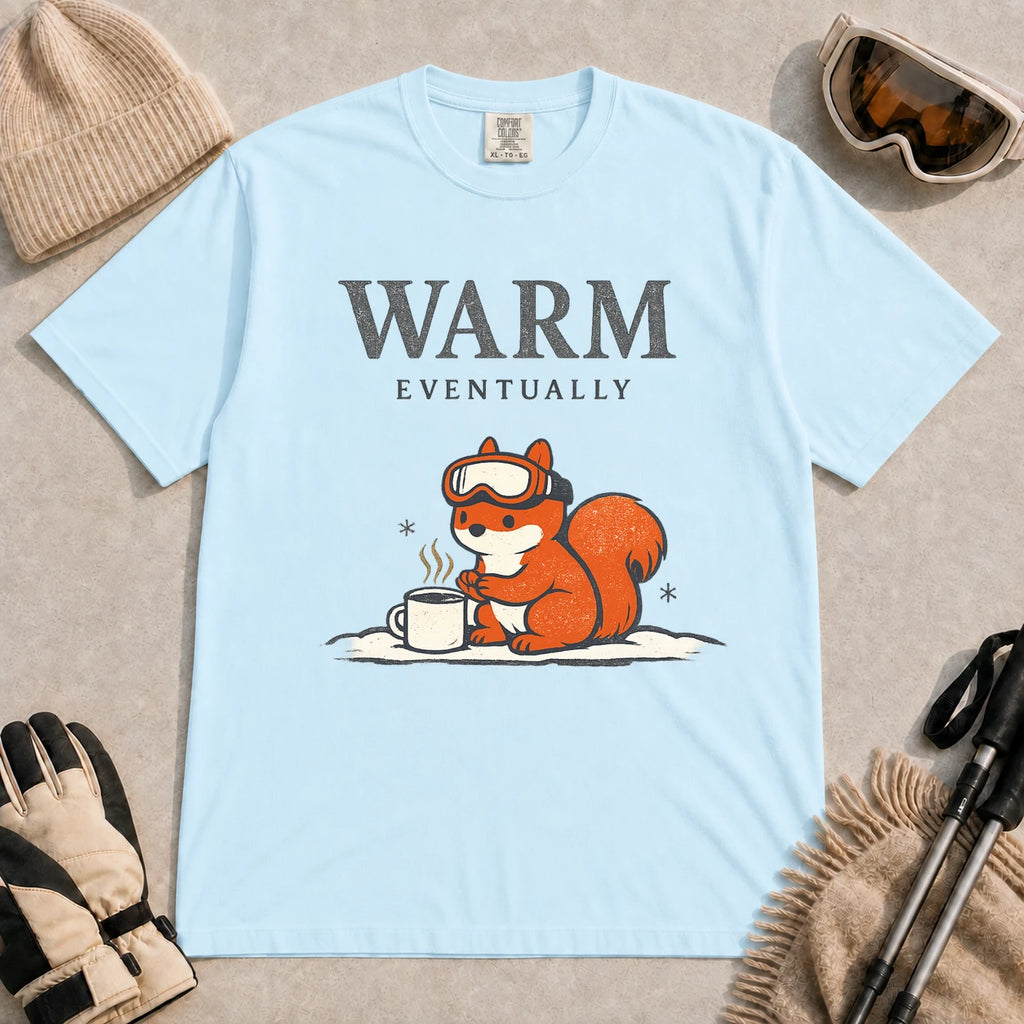 Chambray tee featuring a chilly squirrel in ski goggles warming its paws beside a steaming mug in the snow, with the phrase “Warm Eventually.”
