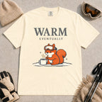 Ivory tee featuring a chilly squirrel in ski goggles warming its paws beside a steaming mug in the snow, with the phrase “Warm Eventually.”