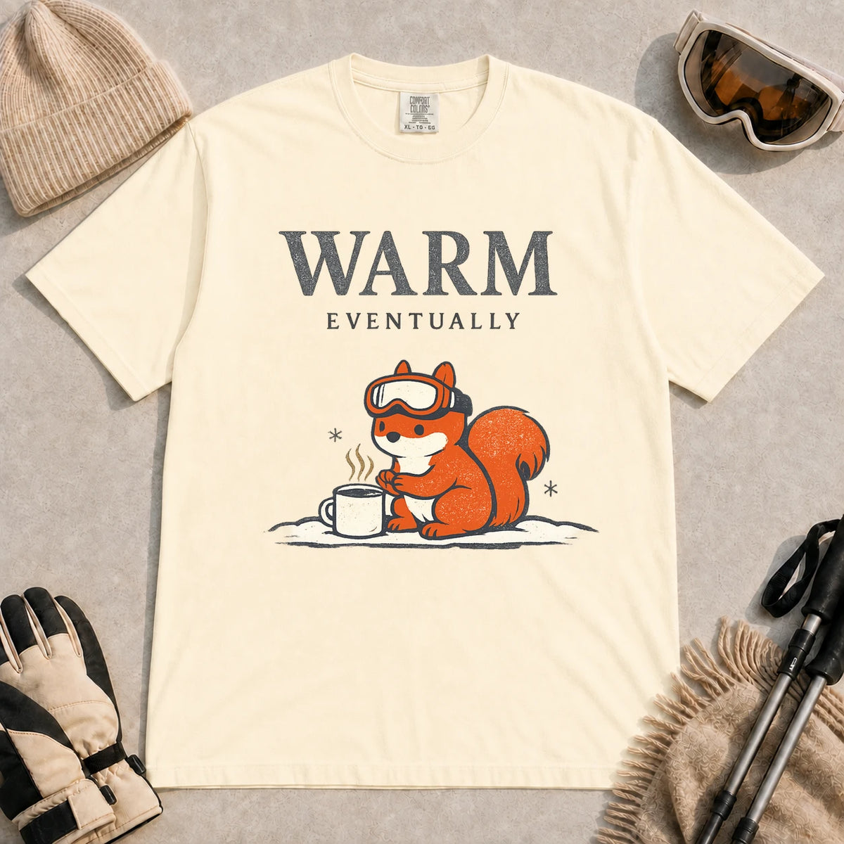 Ivory tee featuring a chilly squirrel in ski goggles warming its paws beside a steaming mug in the snow, with the phrase “Warm Eventually.”
