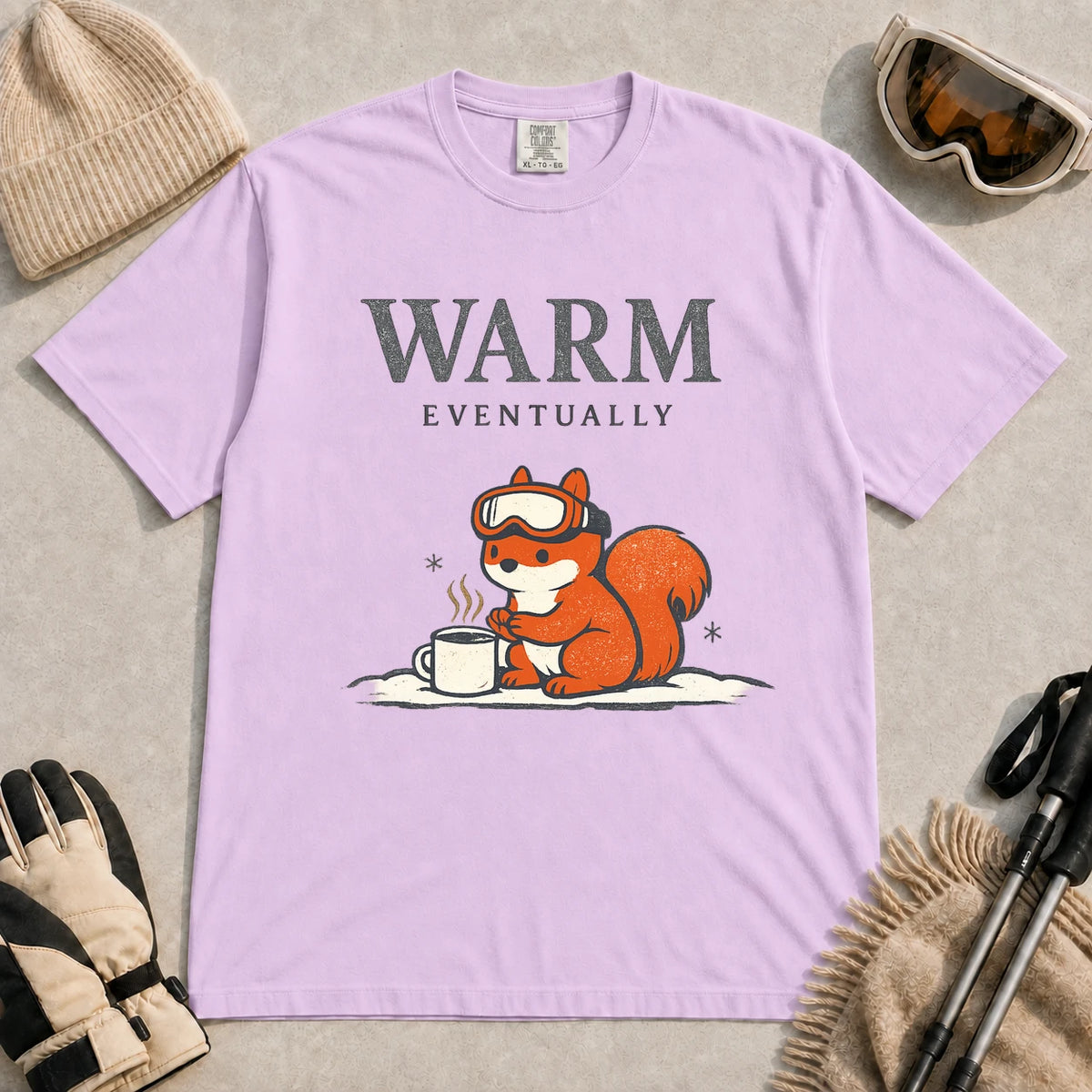 Orchid tee featuring a chilly squirrel in ski goggles warming its paws beside a steaming mug in the snow, with the phrase “Warm Eventually.”