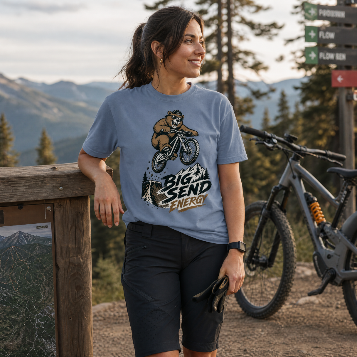 Woman wearing a blue jean colored t-shirt with a bear graphic and text, standing next to a mountain bike with a scenic background.
