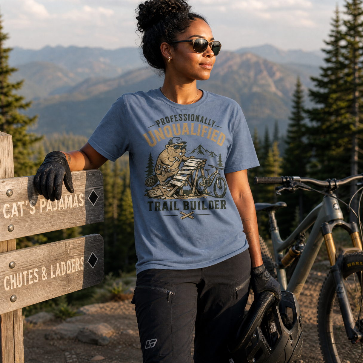 Woman in a blue t-shirt with a mountain bike graphic, standing next to a bike in a forested area with mountains in the background.