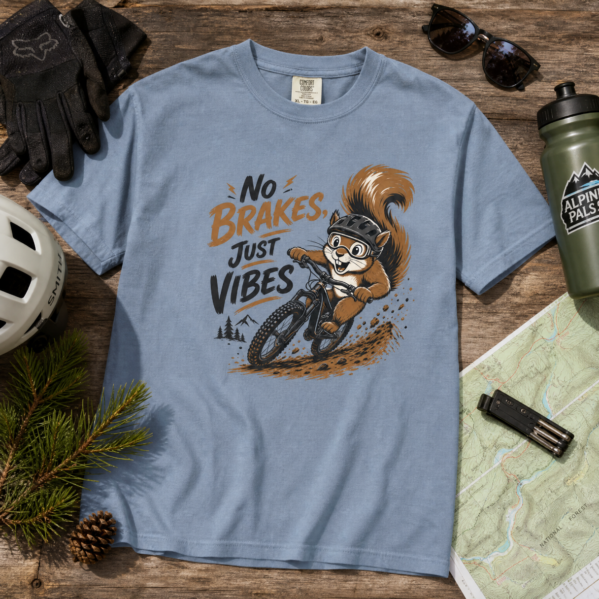 Blue t-shirt with a squirrel graphic and 'No Brakes, Just Vibes' text on a wooden surface with outdoor gear.