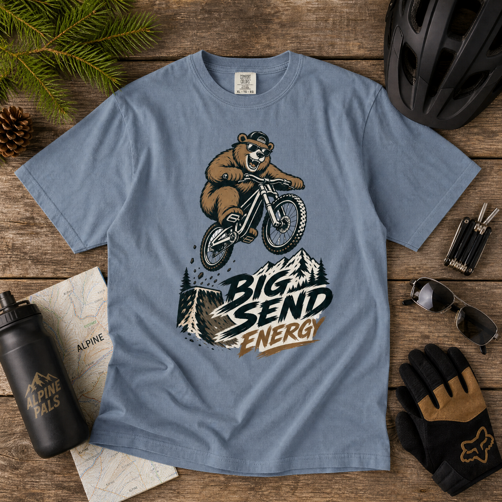 Blue jean t-shirt with bear riding a bike graphic and 'Big Send Energy' text on a wooden surface with cycling accessories.
