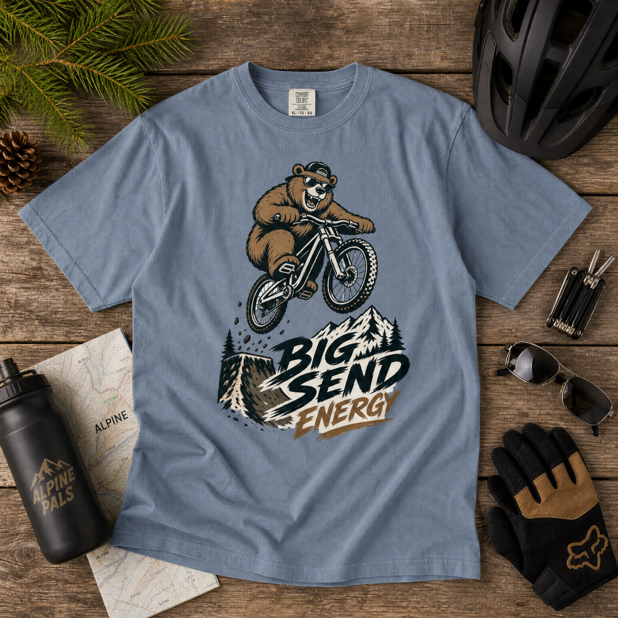 Blue jean t-shirt with bear riding a bike graphic and 'Big Send Energy' text on a wooden surface with cycling accessories.