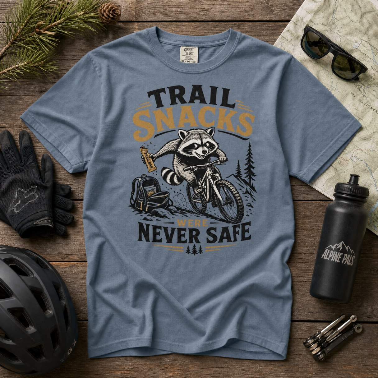Blue jean colored t-shirt with raccoon graphic and text on a wooden surface with outdoor gear.