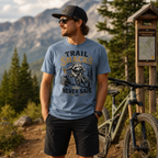 Man wearing a 'Trail Snacks Never Safe' t-shirt in a mountainous area with a bike.