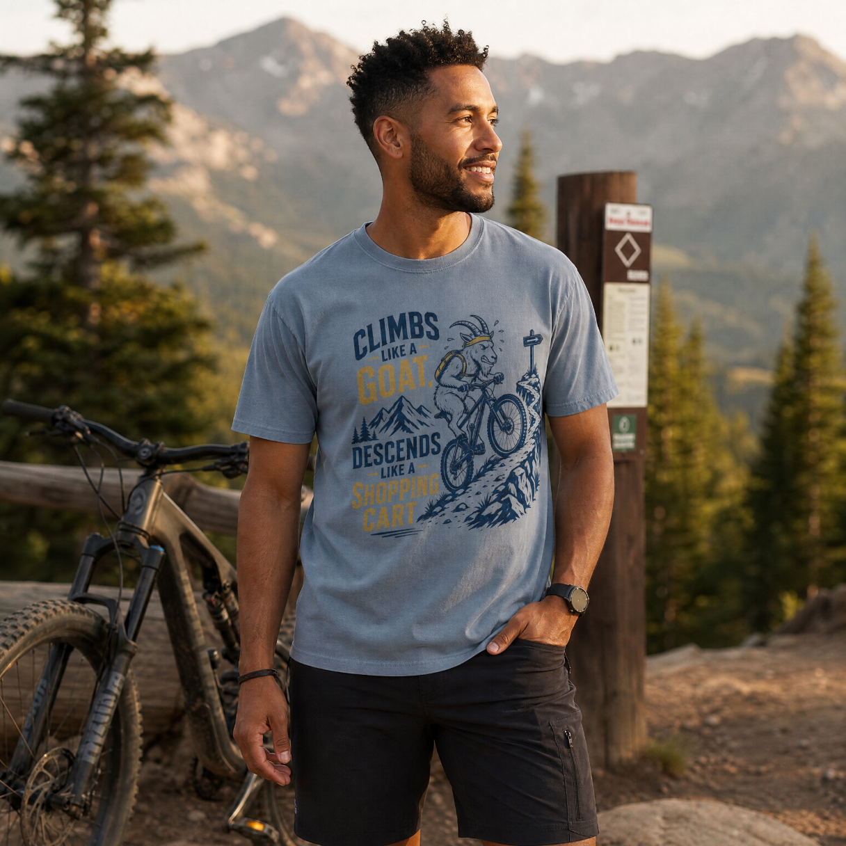 Man wearing a t-shirt with mountain bike design and text, standing in a mountainous area with a bicycle.
