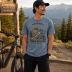 Man wearing a t-shirt with a bear graphic and text, standing next to a bicycle with a mountainous background.