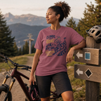 Woman in a Brick colored t-shirt with mountain bike graphic standing next to a bicycle on a trail with mountains in the background.