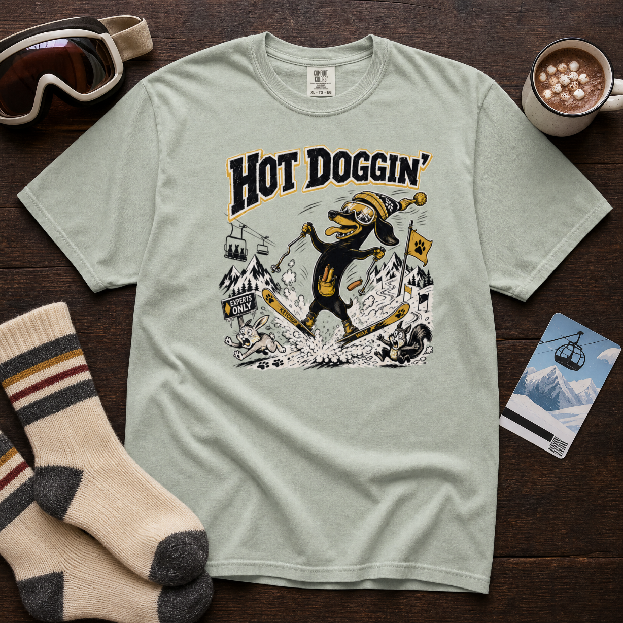 Light green t-shirt with 'Hot Doggin' graphic design on a wooden surface with accessories.