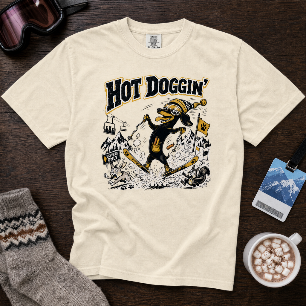 Beige t-shirt with a graphic design of a dog skiing and text 'Hot Doggin'' on a wooden surface with accessories.