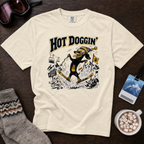 Beige t-shirt with a graphic design of a dog skiing and text 'Hot Doggin'' on a wooden surface with accessories.