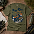 Green t-shirt with fox graphic and 'Flow State Activated' text on a wooden surface with outdoor gear.