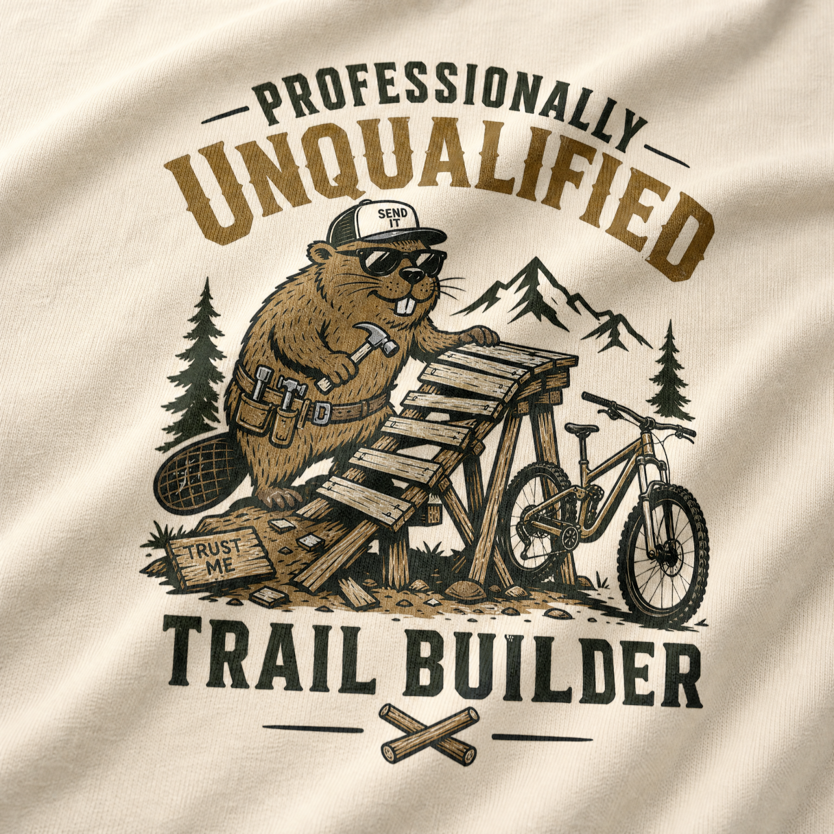 Beige t-shirt with a graphic of a beaver building a trail with text 'Professionally Unqualified Trail Builder' on a mountainous background.
