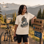 Woman standing next to a bicycle with a mountain bike graphic t-shirt, surrounded by trees and mountains.