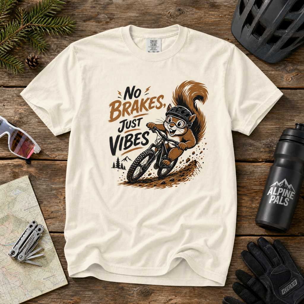 White t-shirt with a graphic of a squirrel riding a bike and 'No Brakes, Just Vibes' text on a wooden surface with cycling accessories.