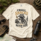 Ivory colored t-shirt with raccoon graphic and 'Trail Snacks Were Never Safe' text on a wooden surface with outdoor gear.